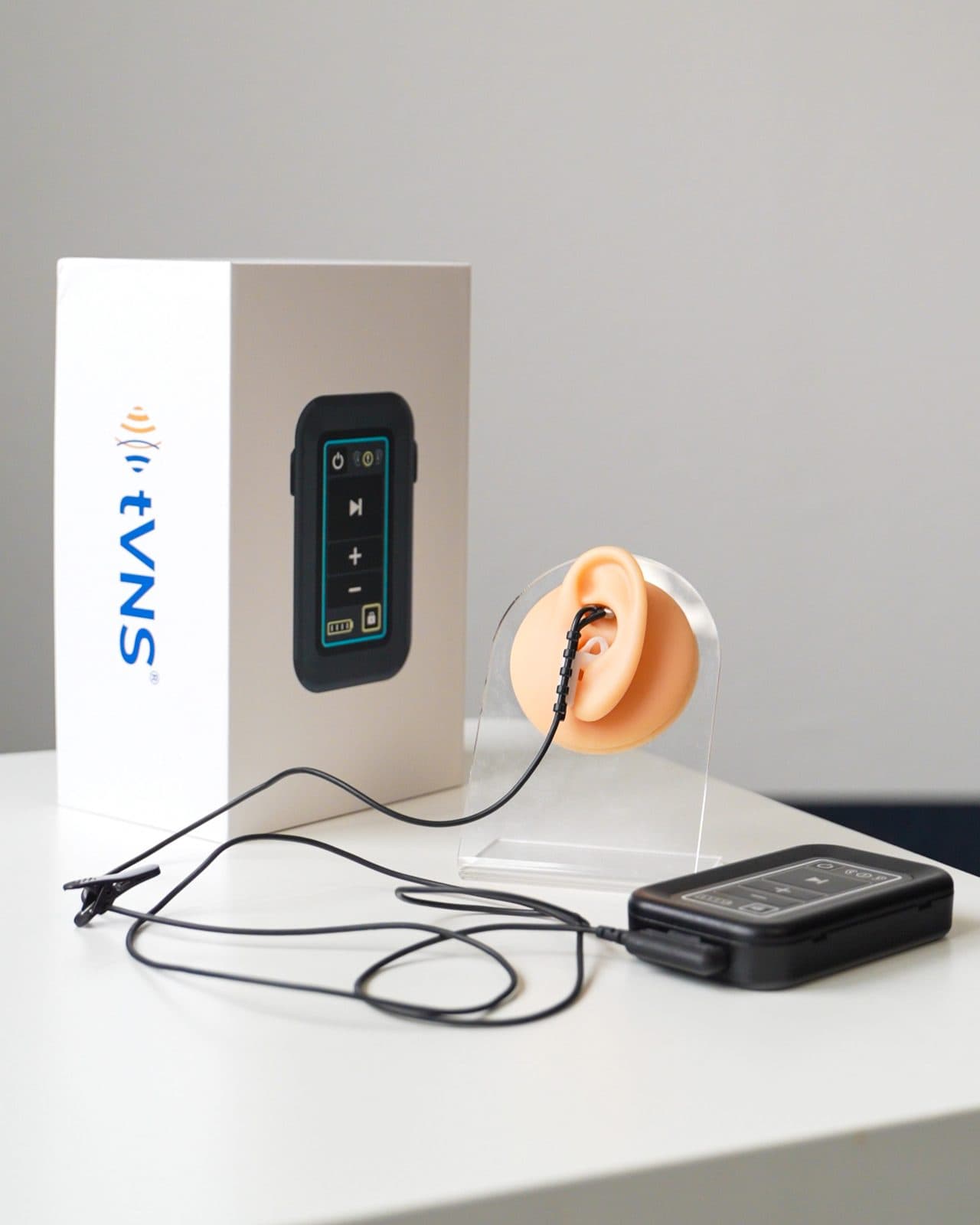 The white tVNS retail box standing beside the stimulator unit, with the ear electrode shown fitted to a display model of an ear.