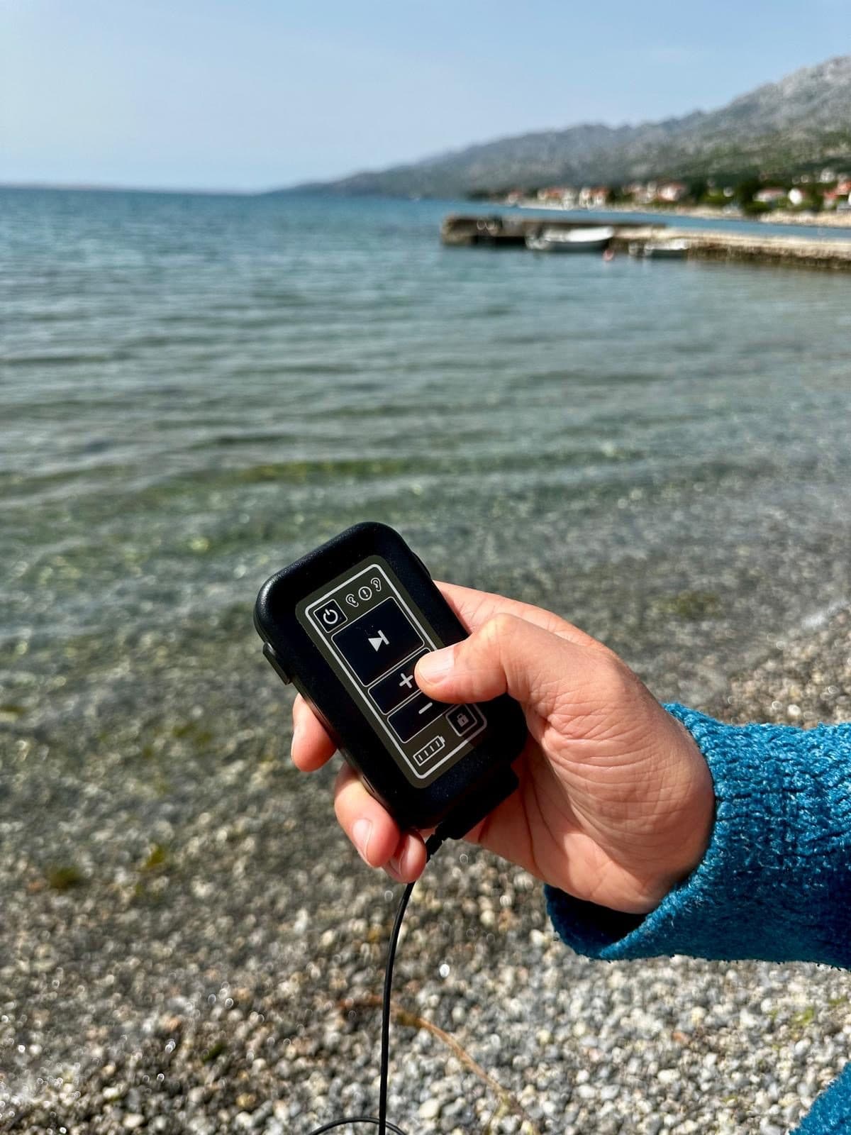 A hand holding the tVNS stimulator outdoors beside the sea, thumb on the intensity controls.